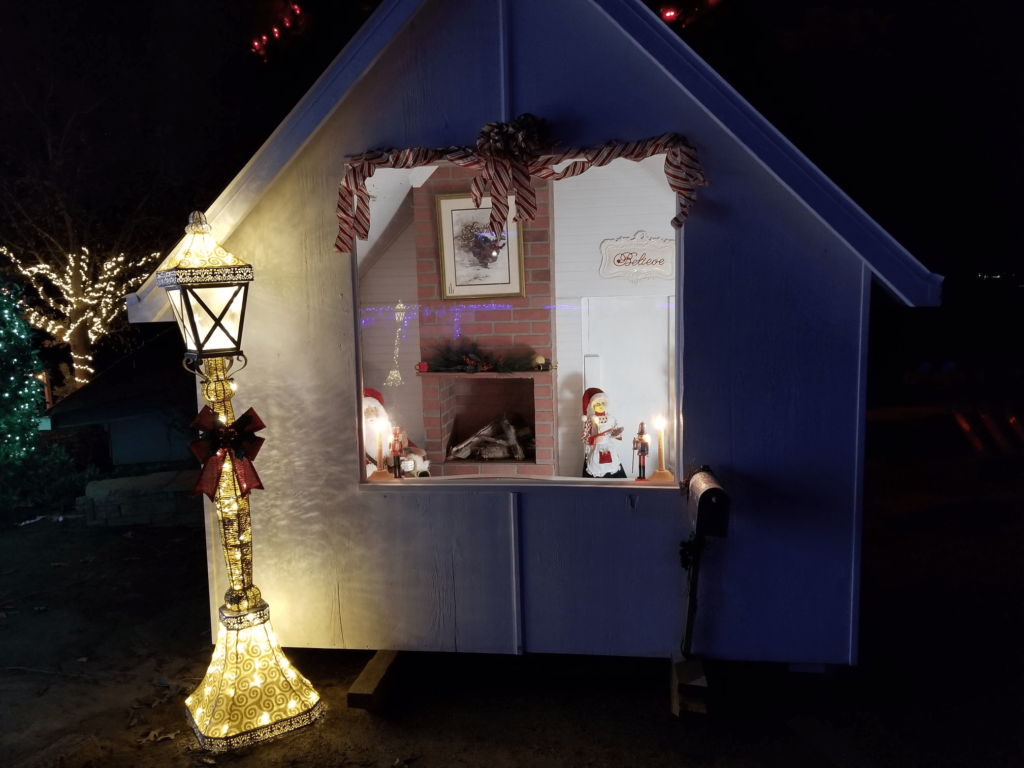 Night scene of a small cabin with festive decorations, including a glowing lamp post, lit window display, and holiday ornaments.