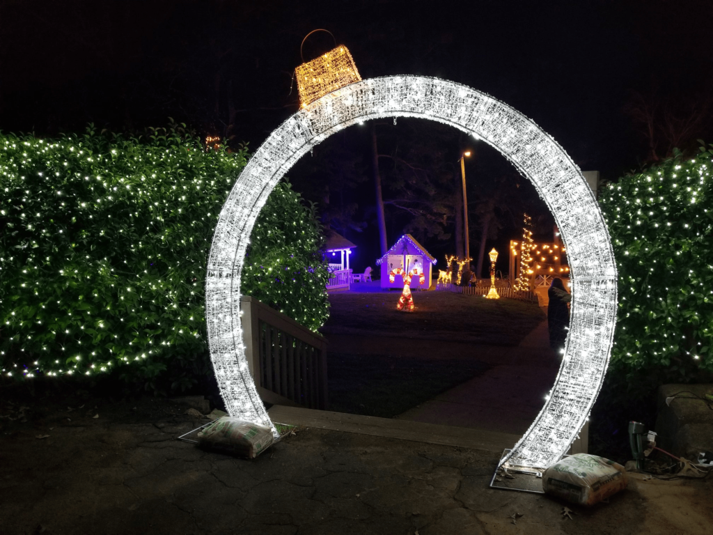 Illuminated archway with Christmas lights leading to a festive garden scene with lit decorations and a small house.